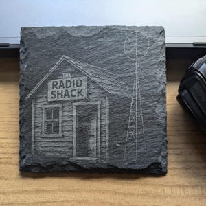 May include: Dark grey square slate coaster featuring an etched design of "The Radio Shack" building and a radio tower. The coaster rests on a wooden surface. A laptop and a black device are in the background.