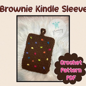 May include: A brown, crocheted Kindle sleeve with colorful, speckled accents. The sleeve has a loop at the top and is designed to resemble a brownie. The text "Brownie Kindle Sleeve" is at the top of the image.