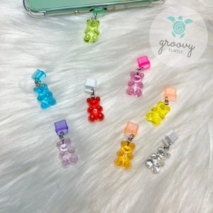 May include: A set of nine gummy bear charms in various colors, including green, blue, pink, red, orange, yellow, and clear. The charms are attached to small metal rings and are designed to be used as book markers or phone charms. The charms are displayed on a white fuzzy surface.