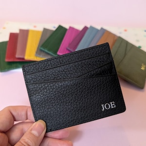May include: A black leather card holder with the name "JOE" in white lettering. Several other colorful card holders are visible in the background. The card holder has multiple card slots and a textured surface. The background is a light pink color.