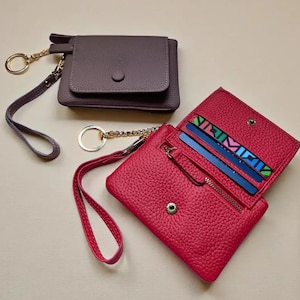 two leather wallet and keychain accessories, one in a dark brown color and the other in a vibrant red color, placed on a light-colored surface.