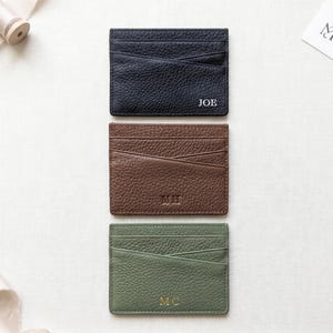 Peut inclure: Trois porte-cartes rectangulaires en cuir noir, marron et vert sauge, avec un grain texturé, plusieurs fentes pour cartes et des initiales personnalisées embossées sur le devant. Le noir affiche JOE, le marron MH et le vert MC.
