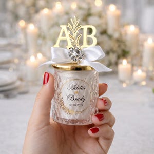 May include: A small, clear glass candle holder with a gold lid and a white ribbon. The candle holder has a diamond pattern and a gold topper with the initials "A" and "B". The candle holder has a label with the names "Adelia Brady" and the date "09.09.2029".