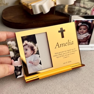 Op de afbeelding: Een goudkleurige fotolijst met de naam "Amelia" en een kruisontwerp. De lijst bevat een foto van een baby en tekst uit 1 Johannes 3:1. Een wit lint met een gouden kruis staat op de achtergrond.