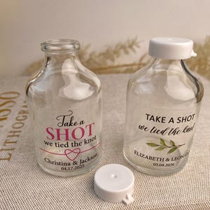 Puede incluir: Dos botellas de vidrio transparente con tapas blancas. Una con el texto "Take a SHOT we tied the knot" y los nombres Christina & Jackson y la fecha 04.17.2025. La otra tiene el texto "Take a SHOT we tied the knot" y los nombres Elizabeth & Leon y la fecha 05.08.2026.
