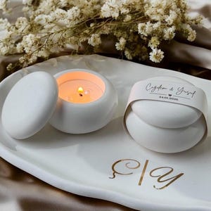 May include: A white ceramic tray with gold lettering reading Ç|y, featuring a lit candle in a round white container with a matching lid, and a second white container wrapped in a paper band printed with Çiğdem & Yusuf and 24.01.2026.