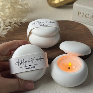 Peut inclure: Des bougeoirs ronds blancs avec couvercles, dont un allum&eacute; avec une flamme. Chacun porte une bande blanche avec les noms "Ashley & Nicholas" et la date 02.06.2026. Les bougeoirs sont sur une surface en bois.