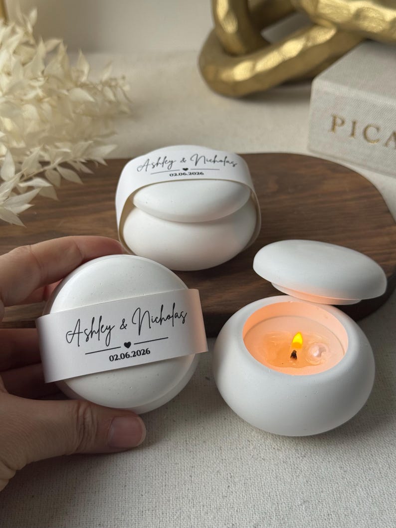Peut inclure: Des bougeoirs ronds blancs avec couvercles, dont un allum&eacute; avec une flamme. Chacun porte une bande blanche avec les noms "Ashley & Nicholas" et la date 02.06.2026. Les bougeoirs sont sur une surface en bois.