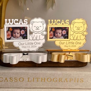 Puede incluir: Dos marcos de fotos para bebés con el nombre "LUCAS" encima de cada foto. Cada marco tiene un diseño de león y el texto "Our Little One May 11, 2026". Un marco es plateado, el otro dorado. Debajo hay cintas decorativas.
