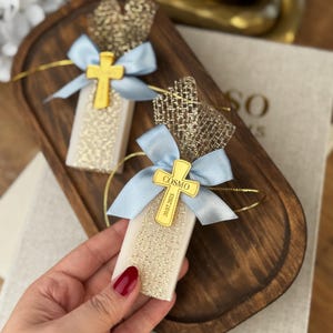 May include: Two rectangular favors with gold glitter, light blue bows, and gold cross charms are displayed on a wooden tray. The charms have the name "COSMO" and the date "20.07.2023" printed on them. The favors are wrapped in gold mesh.