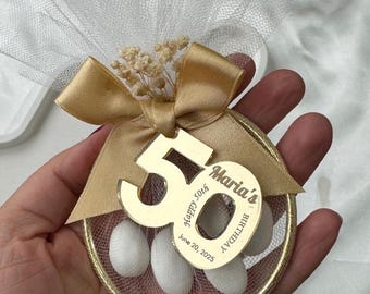 Puede incluir: Un recuerdo dorado con una bolsa de malla llena de caramelos blancos. Una cinta dorada y una etiqueta con el número 50 y el texto "Maria's Birthday June 20, 2025" están adjuntas. El recuerdo está decorado con pequeñas flores secas.