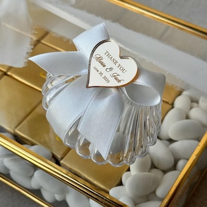 Könnte beinhalten: Eine durchsichtige Plastik-Geschenkbox in Form einer Muschel mit einem weißen Satinband und einem herzförmigen Anhänger mit der Aufschrift "Danke Maria & Jack 20. Juni 2025".