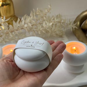 Peut inclure: Bougie blanche sph&eacute;rique avec une bande de papier imprim&eacute;e avec des noms et une date. La bougie est tenue dans une main, avec d'autres bougies allum&eacute;es en arri&egrave;re-plan. Les bougies sont sur un plateau blanc.