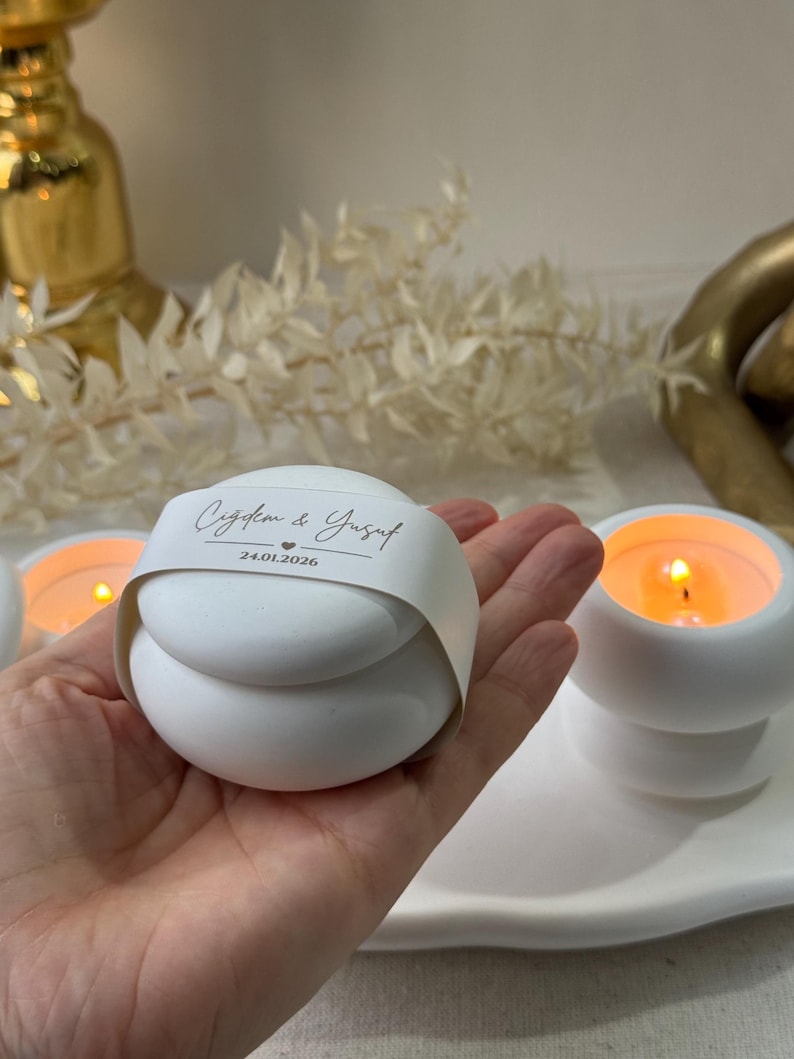 Peut inclure: Bougie blanche sph&eacute;rique avec une bande de papier imprim&eacute;e avec des noms et une date. La bougie est tenue dans une main, avec d'autres bougies allum&eacute;es en arri&egrave;re-plan. Les bougies sont sur un plateau blanc.