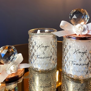 K&ouml;nnte beinhalten: Zwei Glaskerzenhalter mit rautenf&ouml;rmigen Seiten, die jeweils eine wei&szlig;e Kerze enthalten. Die Kerzen sind mit einem Kristallkugel, wei&szlig;em Band und einer wei&szlig;en Blume verziert. Der Text "Xinyu & Tianxiong 05.08.2022" ist aufgedruckt.