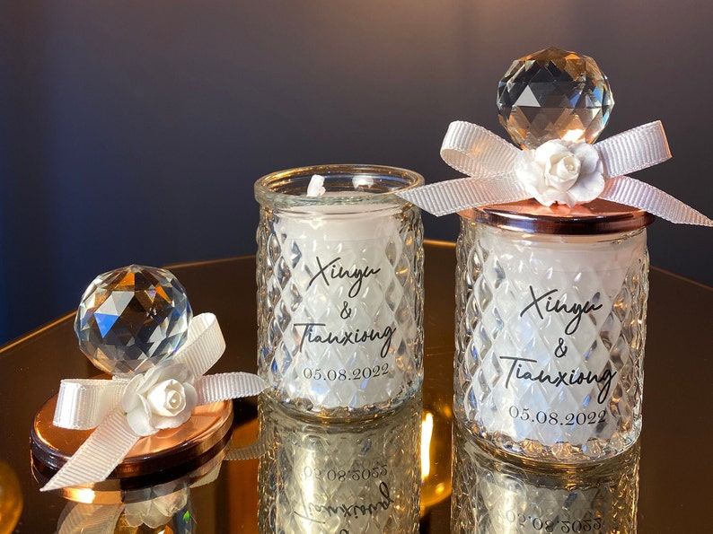K&ouml;nnte beinhalten: Zwei Glaskerzenhalter mit rautenf&ouml;rmigen Seiten, die jeweils eine wei&szlig;e Kerze enthalten. Die Kerzen sind mit einem Kristallkugel, wei&szlig;em Band und einer wei&szlig;en Blume verziert. Der Text "Xinyu & Tianxiong 05.08.2022" ist aufgedruckt.
