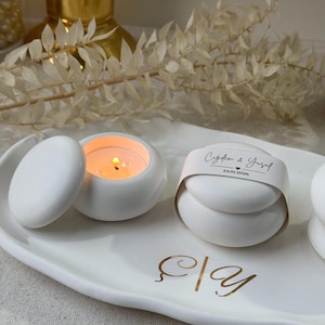 Peut inclure: Bougies en c&eacute;ramique blanches, dont une allum&eacute;e et une ferm&eacute;e avec une &eacute;tiquette, sur un plateau ovale blanc avec des lettres dor&eacute;es. Un objet d&eacute;coratif dor&eacute; est en arri&egrave;re-plan. L'&eacute;tiquette contient du texte et une date.