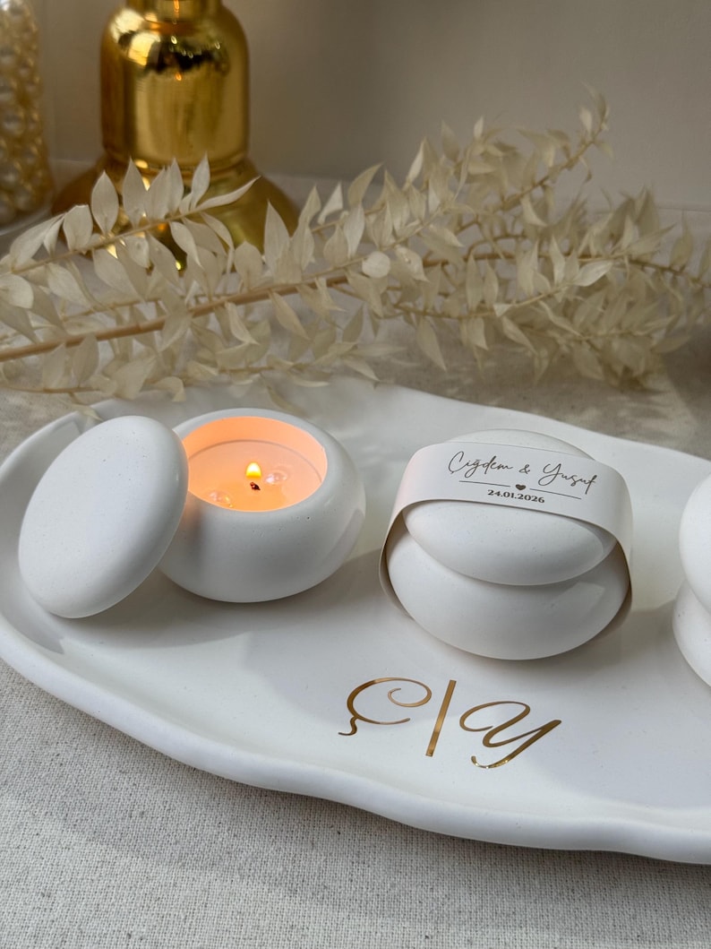 Peut inclure: Bougies en c&eacute;ramique blanches, dont une allum&eacute;e et une ferm&eacute;e avec une &eacute;tiquette, sur un plateau ovale blanc avec des lettres dor&eacute;es. Un objet d&eacute;coratif dor&eacute; est en arri&egrave;re-plan. L'&eacute;tiquette contient du texte et une date.