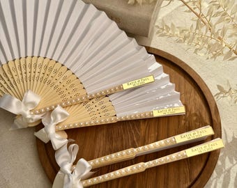 Op de afbeelding: Witte papieren waaiers met houten handvatten, versierd met parels en witte satijnen strikken. Elke waaier heeft een gouden naamplaatje met de tekst "KATIE & PAUL 08.13.2025". De waaiers liggen op een houten dienblad, ideaal voor bruiloften.