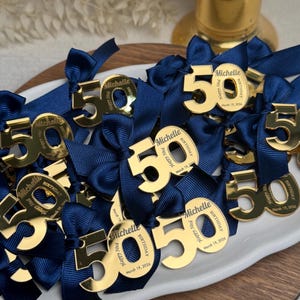 Puede incluir: Una colección de dijes dorados con el número "50", cada uno con una cinta azul marino. Los dijes están personalizados y dispuestos sobre una fuente ovalada blanca, sugiriendo una celebración, posiblemente un cumpleaños.