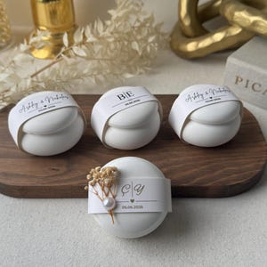 Peut inclure: Quatre objets ronds blancs avec des bandes de papier, dispos&eacute;s sur une planche en bois. Les bandes portent du texte et des dates, l'un des objets &eacute;tant orn&eacute; de fleurs s&eacute;ch&eacute;es et d'une perle. Le fond comprend un d&eacute;cor dor&eacute; et du tissu blanc.