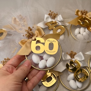 以下が含まれることがあります： 金色の60歳の誕生日記念品。各記念品は、金の数字60、白いチュールが付いた金のリング、白いキャンディーが特徴です。記念品は金色のリボンとドライフラワーで飾られています。「Amanda's 60th Birthday」の文字が見えます。