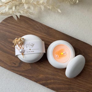 Peut inclure: Deux bougies blanches et rondes avec une perle et une d&eacute;coration de fleurs s&eacute;ch&eacute;es. Une bougie porte une &eacute;tiquette avec des initiales et la date 06.06.2026. L'autre bougie a une lueur orange chaude. Les bougies sont sur un plateau en bois.