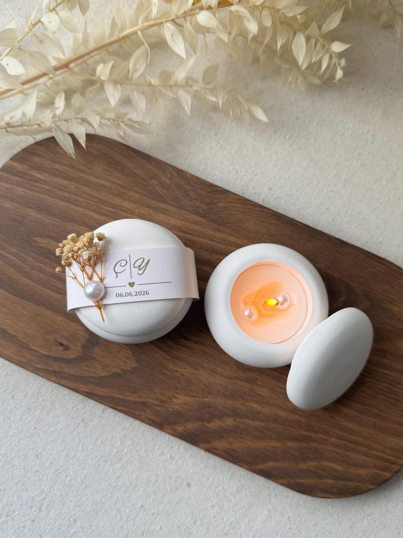 Peut inclure: Deux bougies blanches et rondes avec une perle et une d&eacute;coration de fleurs s&eacute;ch&eacute;es. Une bougie porte une &eacute;tiquette avec des initiales et la date 06.06.2026. L'autre bougie a une lueur orange chaude. Les bougies sont sur un plateau en bois.