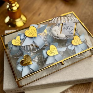 May include: A clear glass box with a gold frame contains several small, clear, fluted containers tied with light blue ribbons. Each container has a gold heart-shaped tag. The box also holds a shell-shaped container with white candies and a card that reads "Thank You, MG & J, August 3, 2023."