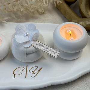 Peut inclure: Des bougeoirs ronds blancs avec des bougies allum&eacute;es, d&eacute;cor&eacute;s de fleurs et de rubans blancs. Une petite &eacute;tiquette avec du texte est attach&eacute;e &agrave; l'un d'eux. Les bougies sont sur un plateau blanc avec des lettres dor&eacute;es.