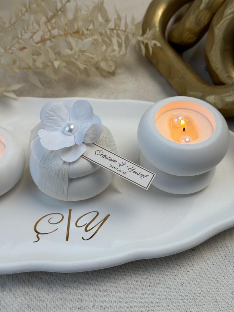 Peut inclure: Des bougeoirs ronds blancs avec des bougies allum&eacute;es, d&eacute;cor&eacute;s de fleurs et de rubans blancs. Une petite &eacute;tiquette avec du texte est attach&eacute;e &agrave; l'un d'eux. Les bougies sont sur un plateau blanc avec des lettres dor&eacute;es.