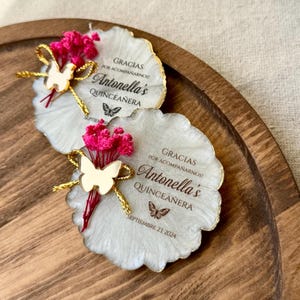 May include: Two white resin coasters with gold accents and a butterfly design. The coasters have the text "Gracias por acompañarnos! Antonella's Quinceañera" and "Septiembre 21, 2024" printed on them. Each coaster is decorated with a small bouquet of pink dried flowers and a gold butterfly.