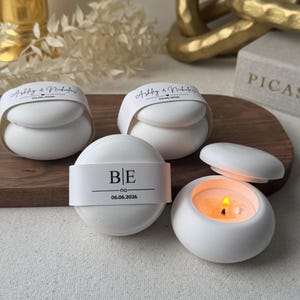 Peut inclure: Des bougeoirs ronds en c&eacute;ramique blanche, dont un avec une bougie allum&eacute;e. Les bougeoirs ont des bandes de papier avec les initiales "BE" et la date "06.06.2026", et les noms "Ashley & Nicholas". Les bougeoirs sont sur une surface en bois.