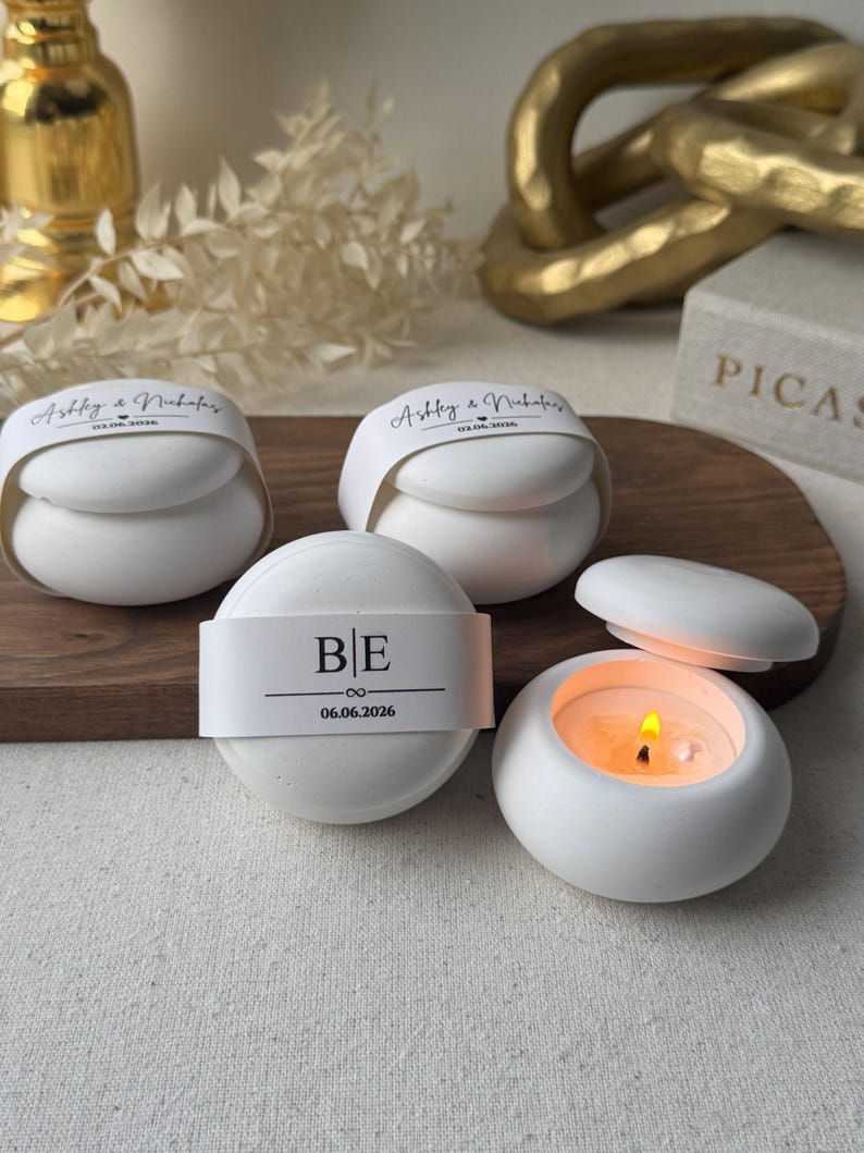 Peut inclure: Des bougeoirs ronds en c&eacute;ramique blanche, dont un avec une bougie allum&eacute;e. Les bougeoirs ont des bandes de papier avec les initiales "BE" et la date "06.06.2026", et les noms "Ashley & Nicholas". Les bougeoirs sont sur une surface en bois.