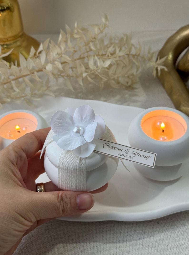 Peut inclure: Bougeoirs blancs avec des bougies allum&eacute;es. Un bougeoir est d&eacute;cor&eacute; d'une fleur blanche et d'un ruban. Une petite &eacute;tiquette indique "&Ccedil;iğdem & Yusuf 24.01.2026". Les bougies sont sur un plateau blanc.