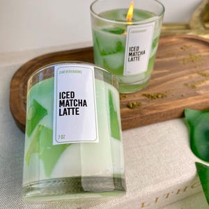 Puede incluir: Dos velas de vidrio con un diseño en capas verde y blanco, etiquetadas como "ICED MATCHA LATTE". Una vela está encendida, mostrando una llama. Las velas pesan 198 gramos cada una. La marca "JJ4EVERFAVORS" también es visible.