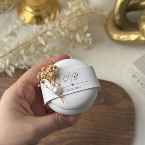 Peut inclure: Une bo&icirc;te &agrave; cadeaux ronde blanche avec une bande d&eacute;corative portant des initiales et une date. Orn&eacute;e de fleurs s&eacute;ch&eacute;es et d'une perle, elle est tenue dans une main. Id&eacute;ale pour un mariage ou un &eacute;v&eacute;nement.