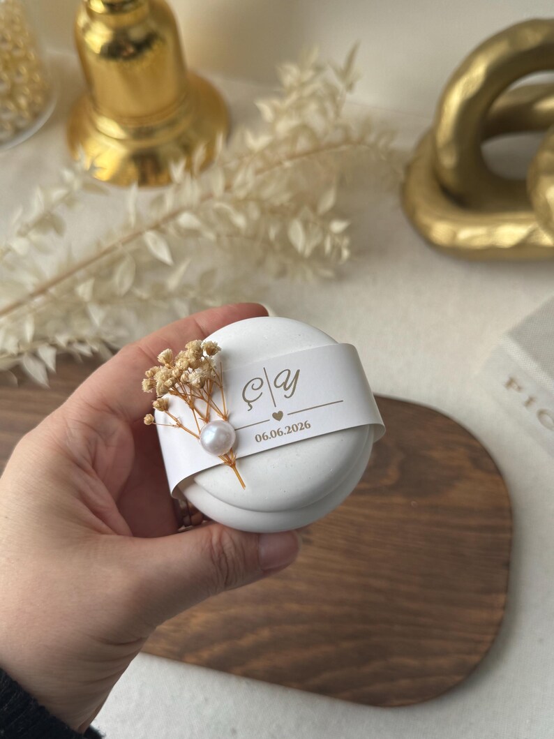 Peut inclure: Une bo&icirc;te &agrave; cadeaux ronde blanche avec une bande d&eacute;corative portant des initiales et une date. Orn&eacute;e de fleurs s&eacute;ch&eacute;es et d'une perle, elle est tenue dans une main. Id&eacute;ale pour un mariage ou un &eacute;v&eacute;nement.