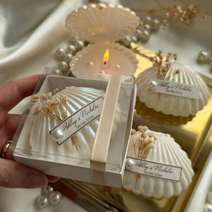 Può includere: Candele bianche a forma di conchiglia con finitura perlata, alcune in scatole trasparenti legate con un nastro. Ogni candela ha un'etichetta con la scritta "Ashley & Nicholas 08/08/2024". Decorate con fiori secchi e perle.