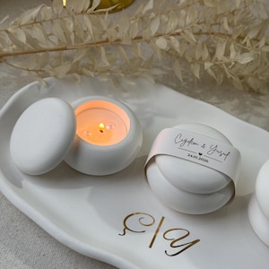 Peut inclure: Un plateau en c&eacute;ramique blanc avec deux petits bougeoirs ronds blancs, dont un avec une bougie allum&eacute;e. Un troisi&egrave;me objet rond est envelopp&eacute; d'une bande de papier avec du texte. &Eacute;criture dor&eacute;e sur le plateau.