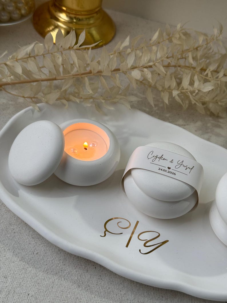 Peut inclure: Un plateau en c&eacute;ramique blanc avec deux petits bougeoirs ronds blancs, dont un avec une bougie allum&eacute;e. Un troisi&egrave;me objet rond est envelopp&eacute; d'une bande de papier avec du texte. &Eacute;criture dor&eacute;e sur le plateau.