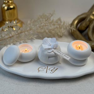 Peut inclure: Un plateau en c&eacute;ramique blanc pr&eacute;sente trois petits bougeoirs blancs avec des bougies allum&eacute;es. L'un a un couvercle, un autre est envelopp&eacute; de tissu et d'une fleur, et le troisi&egrave;me a une &eacute;tiquette. Le plateau a des lettres dor&eacute;es.