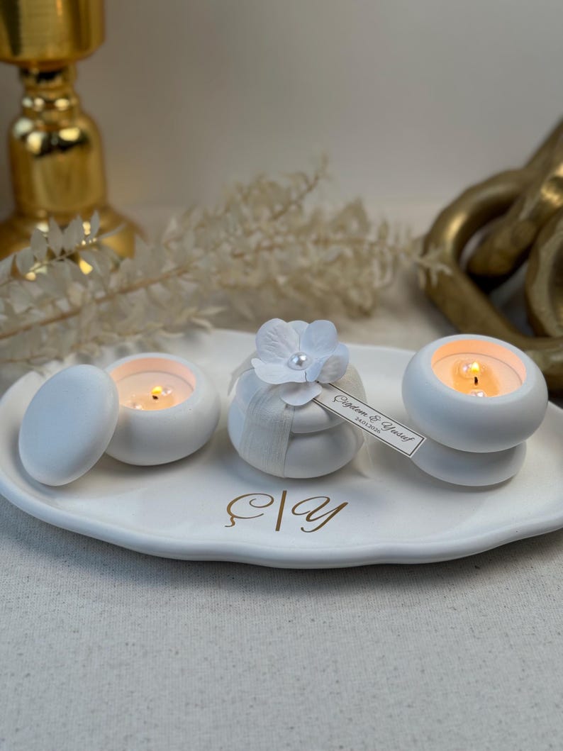 Peut inclure: Un plateau en c&eacute;ramique blanc pr&eacute;sente trois petits bougeoirs blancs avec des bougies allum&eacute;es. L'un a un couvercle, un autre est envelopp&eacute; de tissu et d'une fleur, et le troisi&egrave;me a une &eacute;tiquette. Le plateau a des lettres dor&eacute;es.