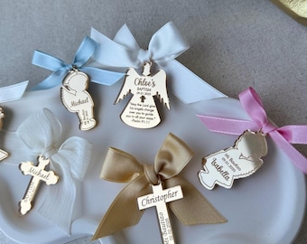 May include: A set of six wooden baptism favors with personalized names and dates. The favors include a cross, an angel, and a child figure. Each favor is tied with a ribbon in a different color.