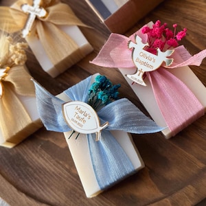 May include: Four rectangular soaps are arranged on a wooden tray. Each soap is decorated with a ribbon and a small embellishment. The ribbons are in shades of gold, blue, and pink. Some soaps have text that reads "Olivia's Baptism" and "Marla's Taufe".