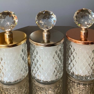 K&ouml;nnte beinhalten: Drei dekorative Glasbeh&auml;lter mit Kristallkugeldeckeln. Die Deckel sind gold-, silber- und kupferfarben. Die Gl&auml;ser haben ein Rautenmuster und enthalten eine wei&szlig;e Substanz. Die Gl&auml;ser stehen auf einer reflektierenden Oberfl&auml;che.