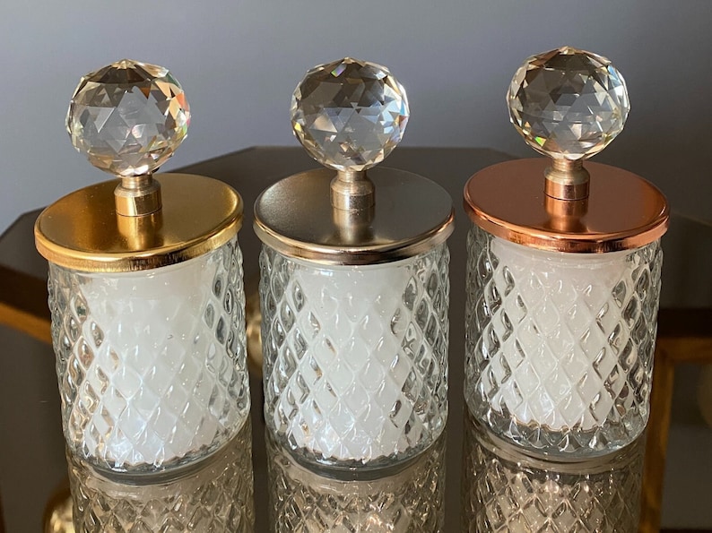 K&ouml;nnte beinhalten: Drei dekorative Glasbeh&auml;lter mit Kristallkugeldeckeln. Die Deckel sind gold-, silber- und kupferfarben. Die Gl&auml;ser haben ein Rautenmuster und enthalten eine wei&szlig;e Substanz. Die Gl&auml;ser stehen auf einer reflektierenden Oberfl&auml;che.