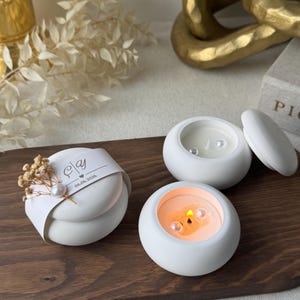 Peut inclure: Trois bougies blanches et rondes avec des perles sont pr&eacute;sent&eacute;es sur un plateau en bois. Une bougie est allum&eacute;e, d&eacute;gageant une lueur chaleureuse. Un article emball&eacute; avec un ruban et des fleurs s&eacute;ch&eacute;es est &eacute;galement pr&eacute;sent.