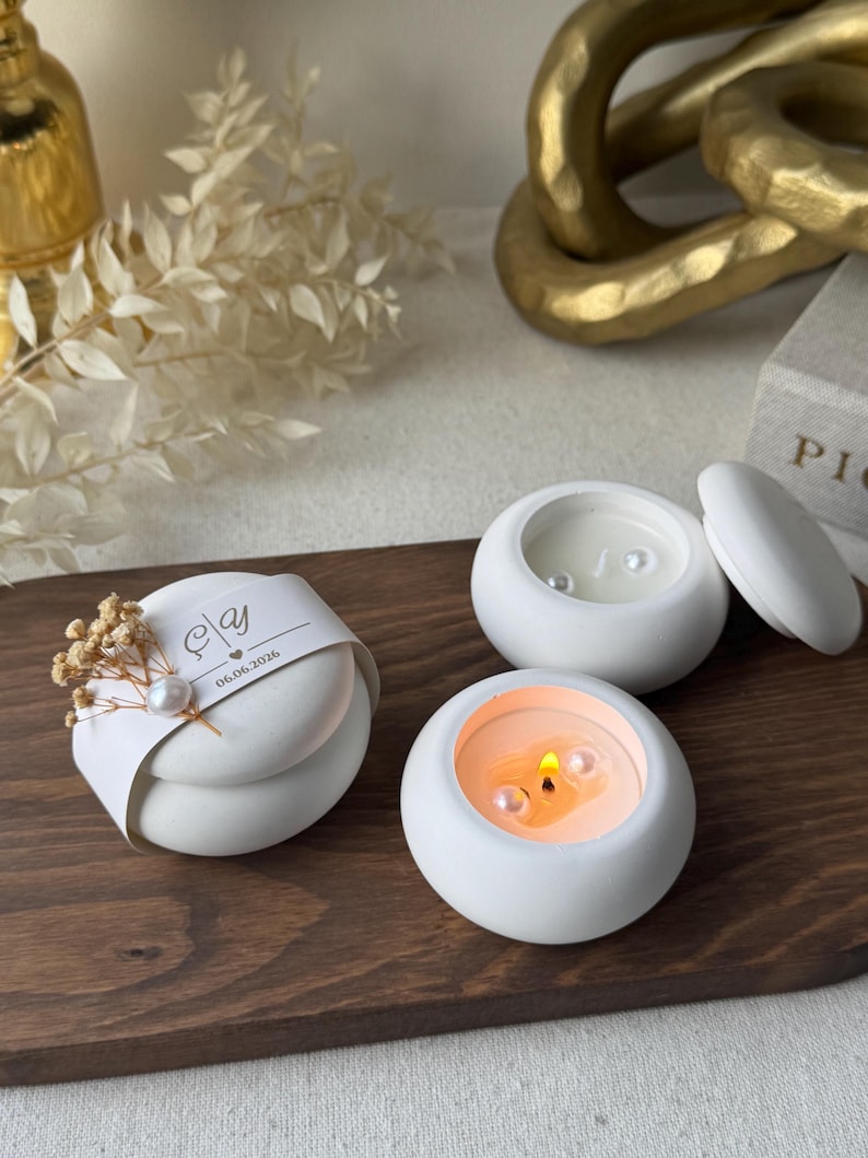 Peut inclure: Trois bougies blanches et rondes avec des perles sont pr&eacute;sent&eacute;es sur un plateau en bois. Une bougie est allum&eacute;e, d&eacute;gageant une lueur chaleureuse. Un article emball&eacute; avec un ruban et des fleurs s&eacute;ch&eacute;es est &eacute;galement pr&eacute;sent.