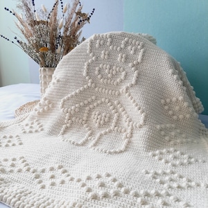 May include: A cream-coloured, handmade crochet blanket with a floral design. The blanket features raised, textured bobbles and a detailed border. The soft, neutral colour and intricate pattern make it a cosy home decor item.